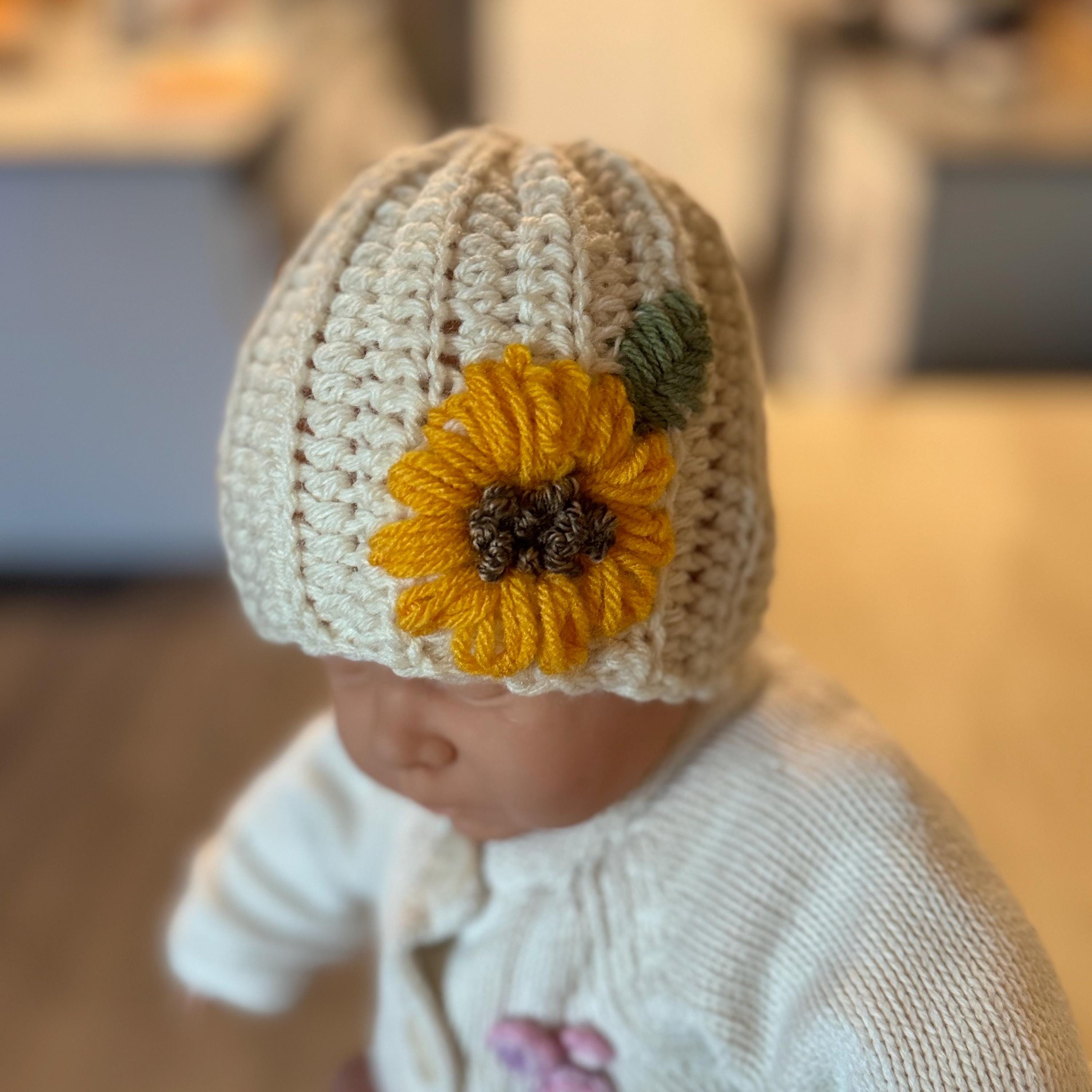 The baby is wearing a crochet beige hat with hand-embroidered Sunflower: yellow petals, brown center, and green leaf.